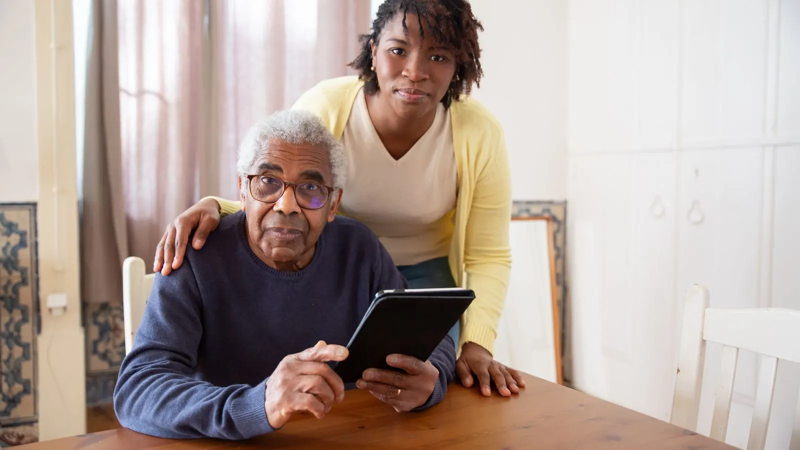 A home caregiver rests her hands warmly on the shoulders of her smiling elderly client at his dining table in Las Vegas